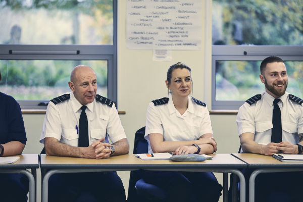 2 male and 1 female prison officers sit in a row in the classroom. On the wall behind is a sheet of paper with illegible words on a 2 windows showing greenery outside