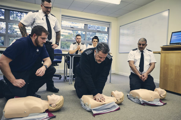 A prison officer simulates CPR on a first aid dummy while 5 others watch. There are 2 other CPR dummies on the floor.