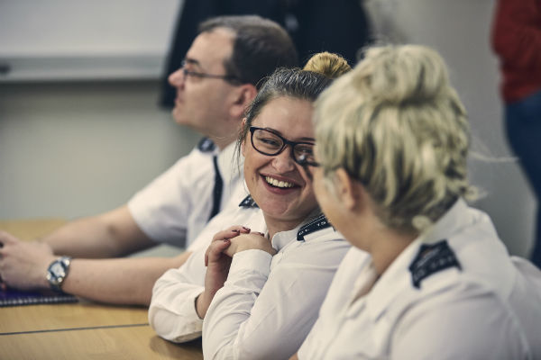 Two female prison officers chatting and smiling while sat behind a desk in a classroom. A third, male officer, sits next to them looking forwards.