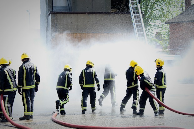 8 people dressed in fire brigade uniform hosing using a fire hose against a wall. 