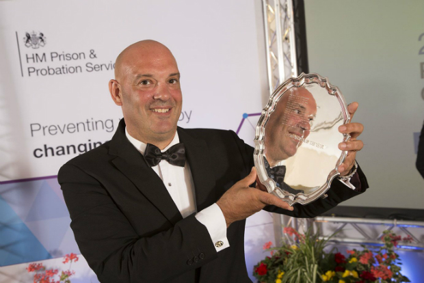 Ian is dressed in a tuxedo and smiling while holding up a silver platter. His face is reflected in the silver and behind him is an HMPPS banner