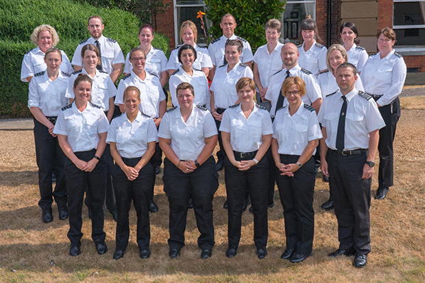 HMPPS Senior Leadership Scheme group picture - 22 men and women stand in 3 lines dressed in black and white prison officer uniforms. They stand outside with a building and hedge behind them