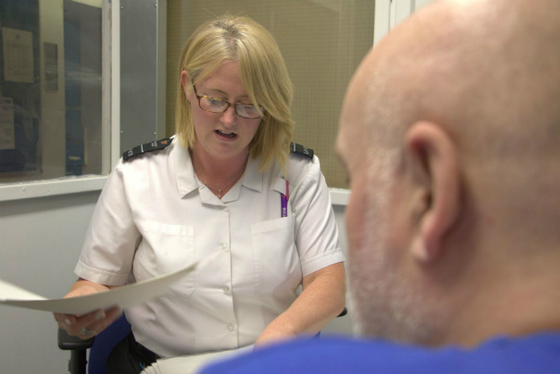 A female prison officer in uniform holds a piece of paper while sat across from a male prisoner. Only the back of the prisoners head is visible.