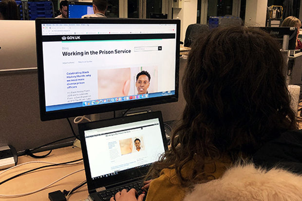 An employee looking at the Working in the Prison Service blog on a computer - the site appears on a laptop screen and on a larger screen above. The employee is sat in an office.