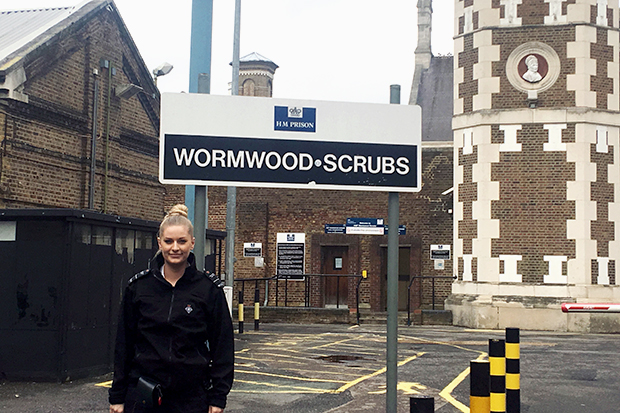 Officer Bowden is dressed in her prison officer uniform outside the prison building. Behind her a large sign says HM Prison Wormwood Scrubs