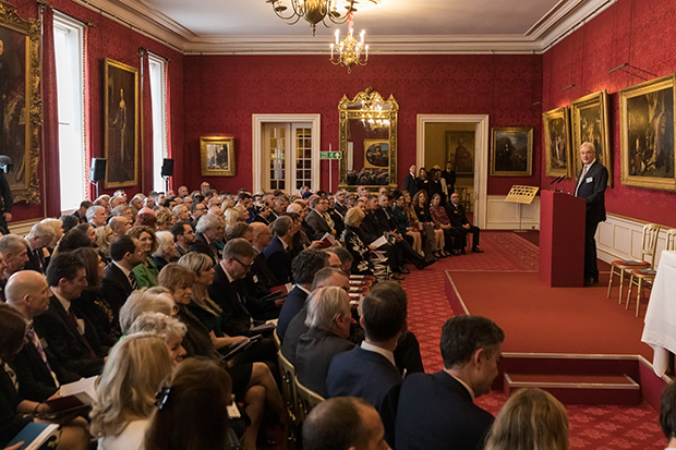 A man stands behind a lectern on a small stage in a large room with red walls and large paintings hanging throughout. The room is filled with an audience of people sat facing the stage.