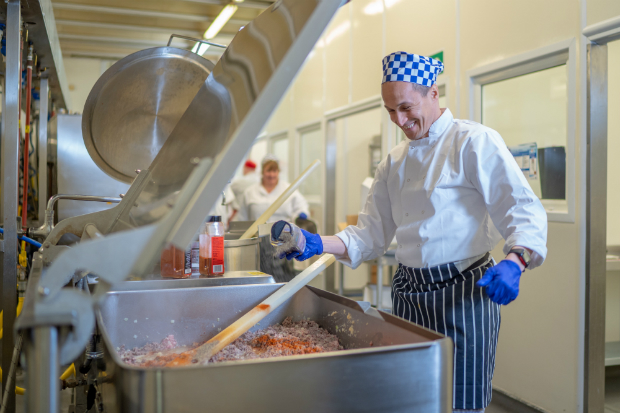 A man dressed in chefs clothing stands over a large metal pan filled with mince and tomatoes with a large wooden spoon. In the background you other people are also cooking.