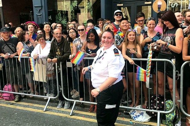 Nic stands in the foreground wearing a black and white uniform. Behind them is a metal barrier and on the other side of that a crowd of people are smiling and waving rainbow flags